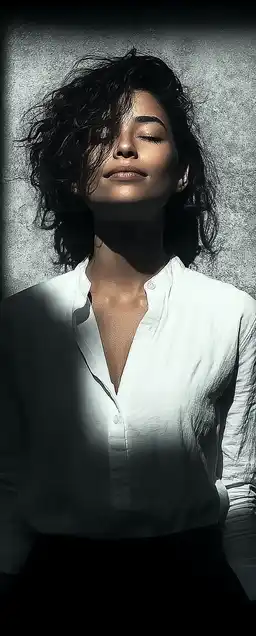 Portrait of a woman wearing a simple white shirt, standing in front of a plain textured wall, natural skin tones and relaxed expression, backlit with clean warm lighting