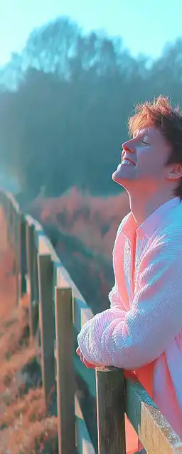A young man leaning against a wooden fence in a countryside field, sleeves rolled up, natural light on his face, captured mid-laugh with trees blurred in the background